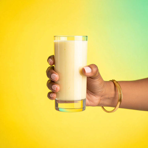 Hand holding a glass of Banana Creme Collagen Keto Shake against a colorful background.