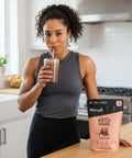 Woman drinking chocolate collagen keto shake in modern kitchen next to product package